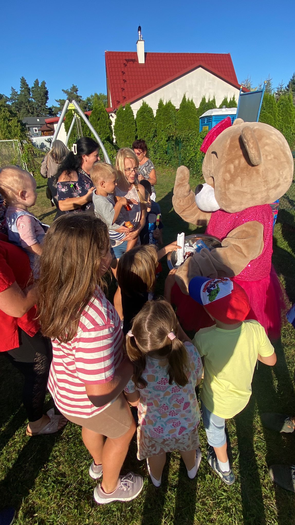 Pluszowy miś w różowej sukience rozdaje słodycze dzieciom na zielonym trawniku, w tle dom z czerwoną dachówką i drzewa.