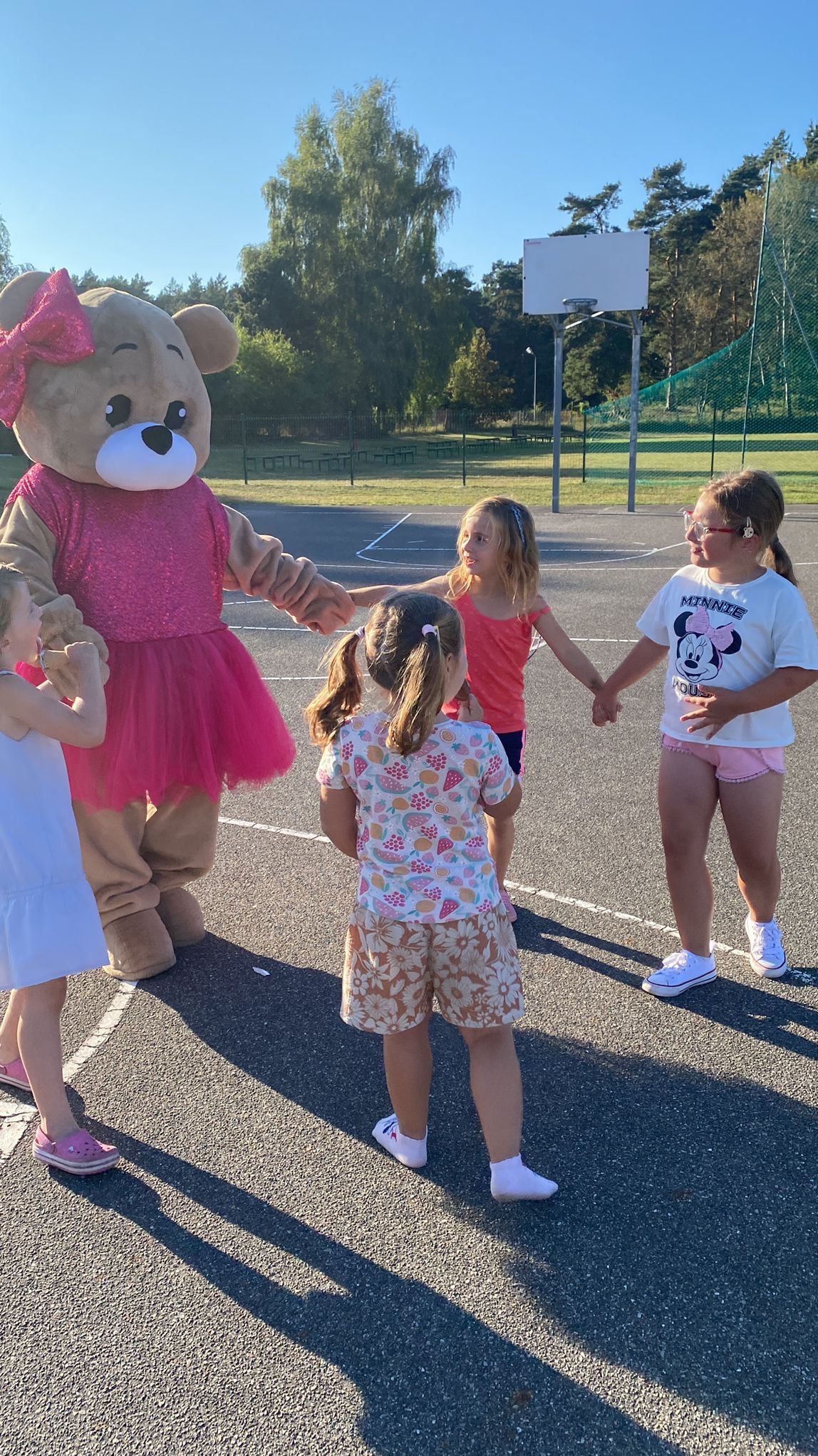 Pluszowy miś w różowej sukience i kokardzie trzyma za ręce grupę dzieci na asfaltowym boisku, w tle drzewa i kosz do koszykówki.