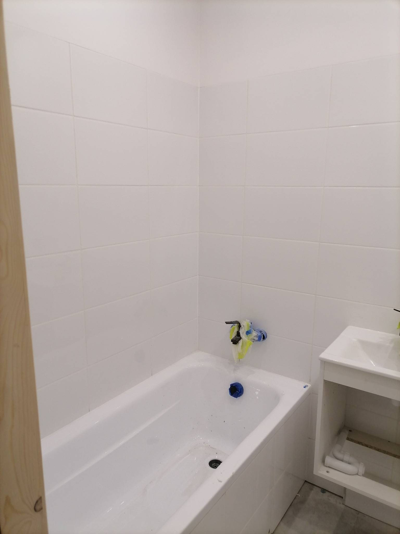 Wnętrze łazienki w trakcie remontu, widoczna biała wanna, umywalka z szafką, białe płytki na ścianach, zabezpieczona bateria i odpływ.