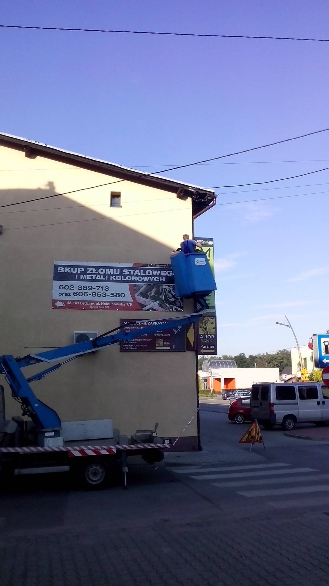 Montaż baneru reklamowego na ścianie budynku za pomocą podnośnika koszowego, widoczny pracownik w niebieskim kombinezonie.