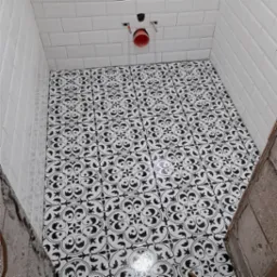 Ułożona podłoga z czarno-białych płytek o wzorze ornamentalnym w małej łazience z białymi, błyszczącymi płytkami ściennymi ułożonymi w cegiełkę, widoczne fragmenty surowych ścian.