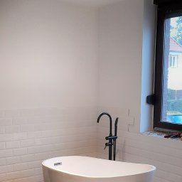 ALLES - Biała, wolnostojąca wanna z czarną armaturą na tle ściany z białych płytek w kształcie cegiełek, oświetlona punktowym światłem