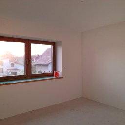 ALLES - Pomalowane na biało wnętrze pokoju z drewnianym oknem, na parapecie stoi czerwone wiaderko.