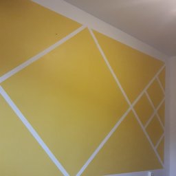 ALLES - Żółta ściana z geometrycznym wzorem z białych linii, świeżo po malowaniu. Widoczne detale wykończenia i gniazdko elektryczne przy podłodze.