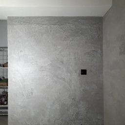 ALLES - Ściana w łazience wykończona mikrocementem w odcieniach szarości, z widocznymi przyłączami wodnymi i gniazdkiem elektrycznym. Minimalistyczny design.