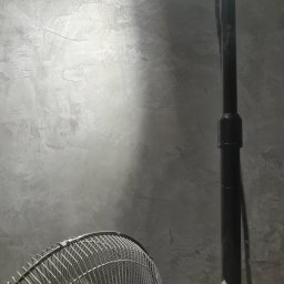 ALLES - Szara ściana z efektem betonu, oświetlona z góry, z fragmentem wentylatora i czarną rurą. Minimalistyczne wykończenie wnętrza.