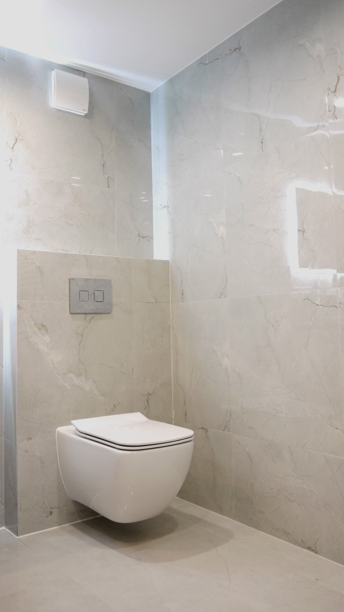 Nowoczesna, podwieszana toaleta w łazience wykończonej płytkami imitującymi marmur, z minimalistycznym panelem spłukującym i wentylatorem sufitowym.