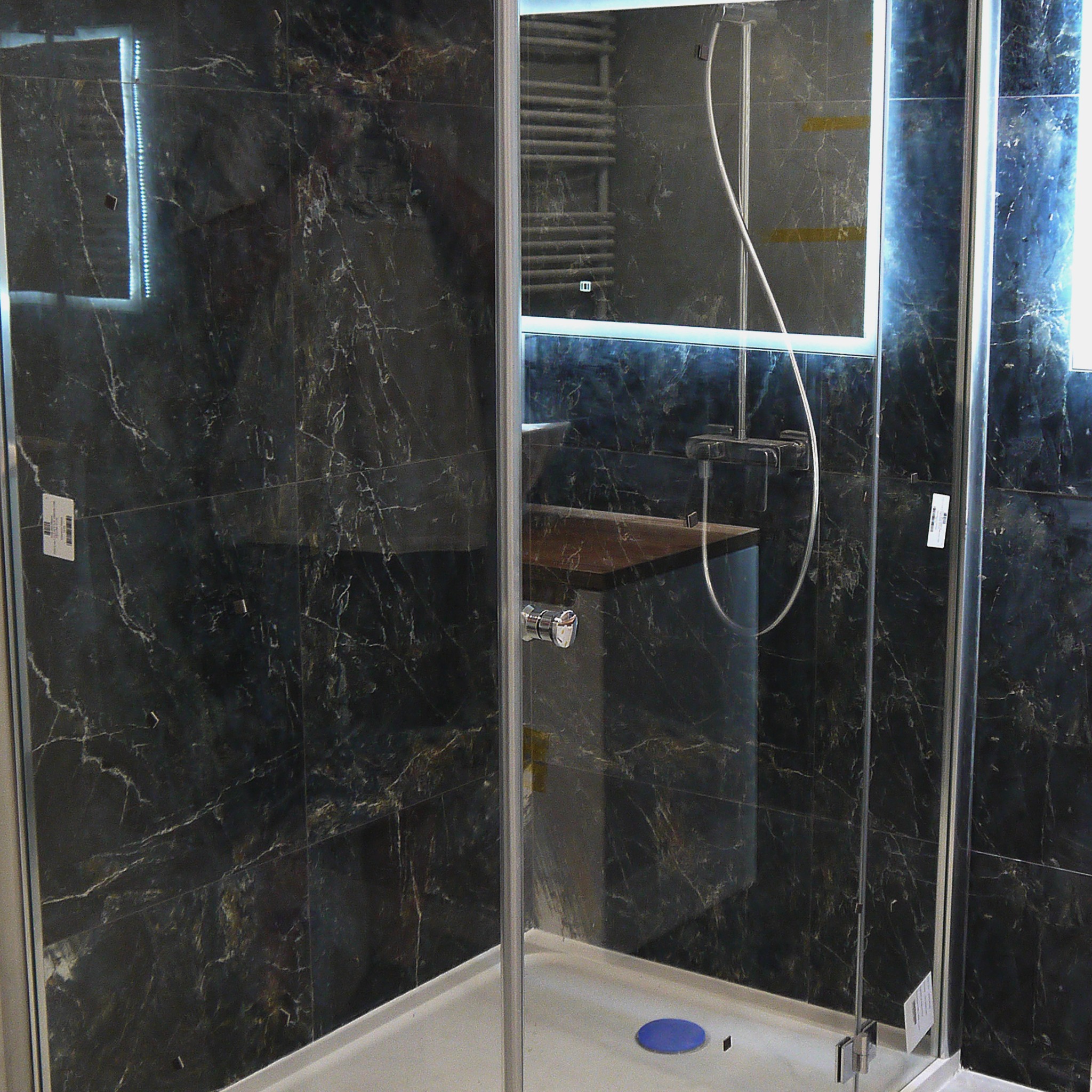 Nowoczesna kabina prysznicowa z ciemnymi marmurkowymi płytkami, podświetlanym lustrem i drewnianym blatem umywalki w minimalistycznej łazience.