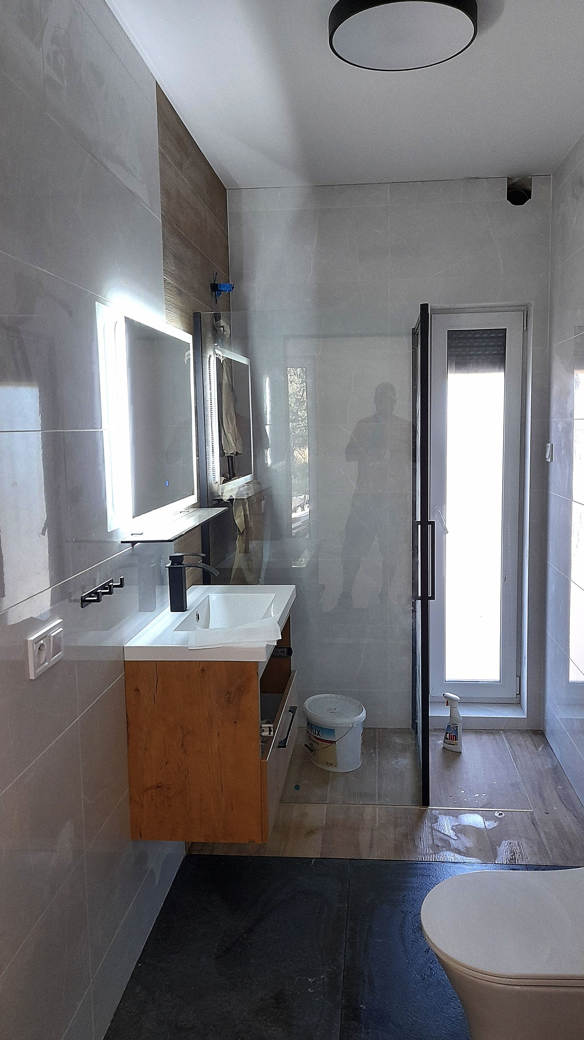 Nowoczesna łazienka z drewnianą szafką pod umywalką, podświetlanym lustrem i kabiną prysznicową typu walk-in, wykończona szarą płytką imitującą kamień i drewnopodobną okładziną na ścianie.