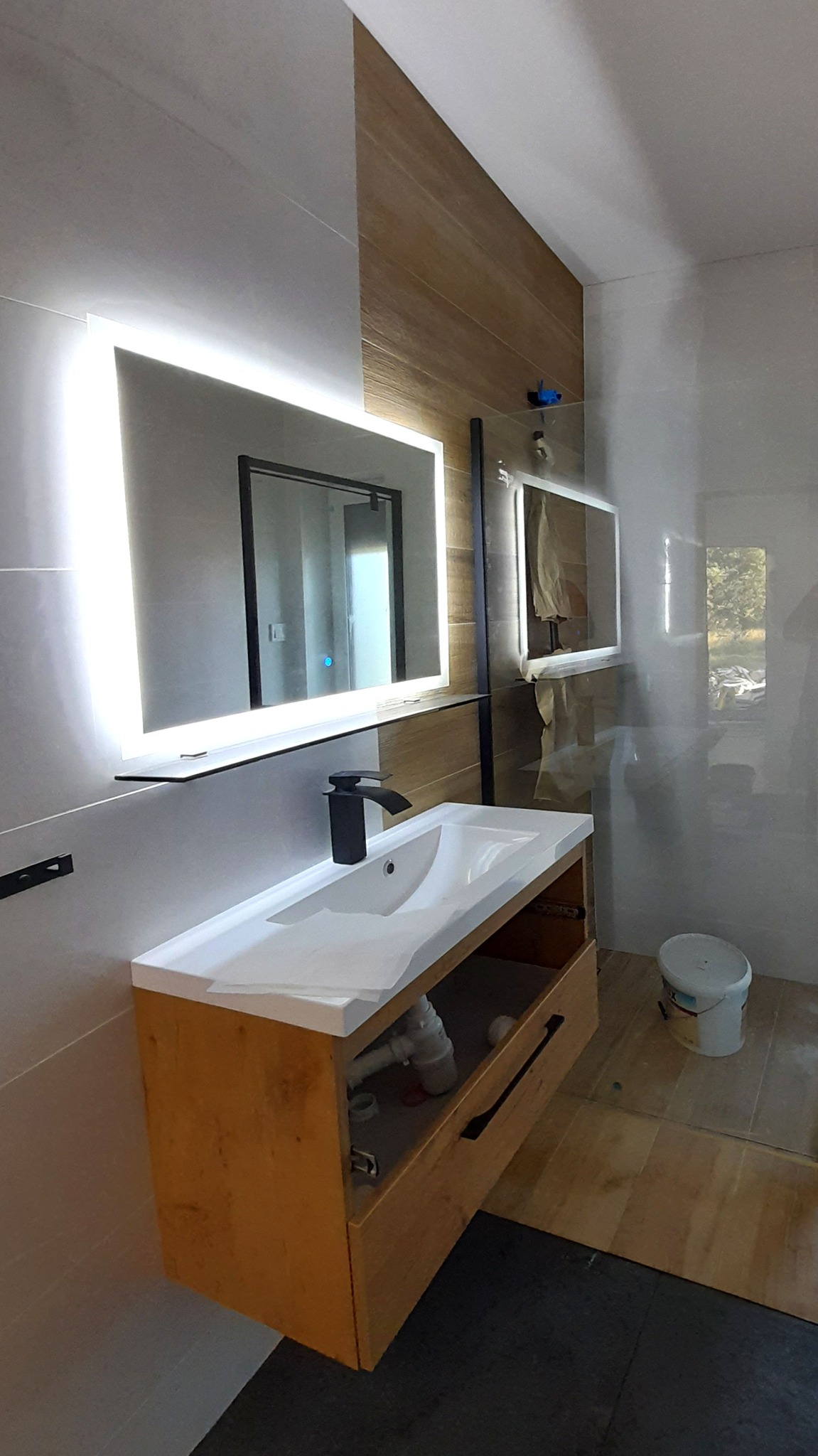 Nowoczesna łazienka w trakcie wykańczania z podświetlanym lustrem, drewnianą szafką pod umywalką i fragmentem niezabezpieczonego lustra na ścianie.