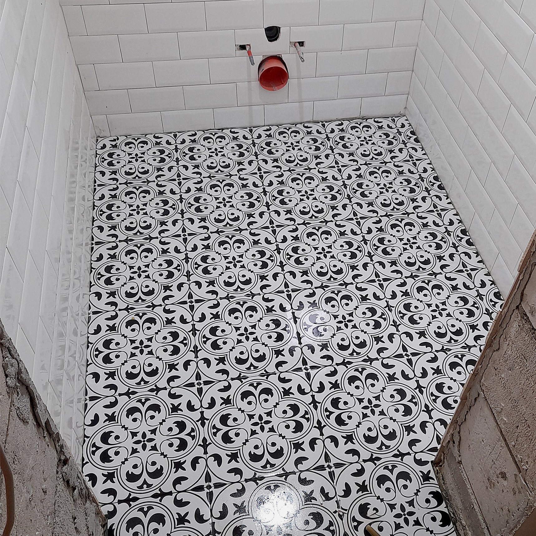 Ułożona podłoga z czarno-białych płytek o wzorze ornamentalnym w małej łazience z białymi, błyszczącymi płytkami ściennymi ułożonymi w cegiełkę, widoczne fragmenty surowych ścian.