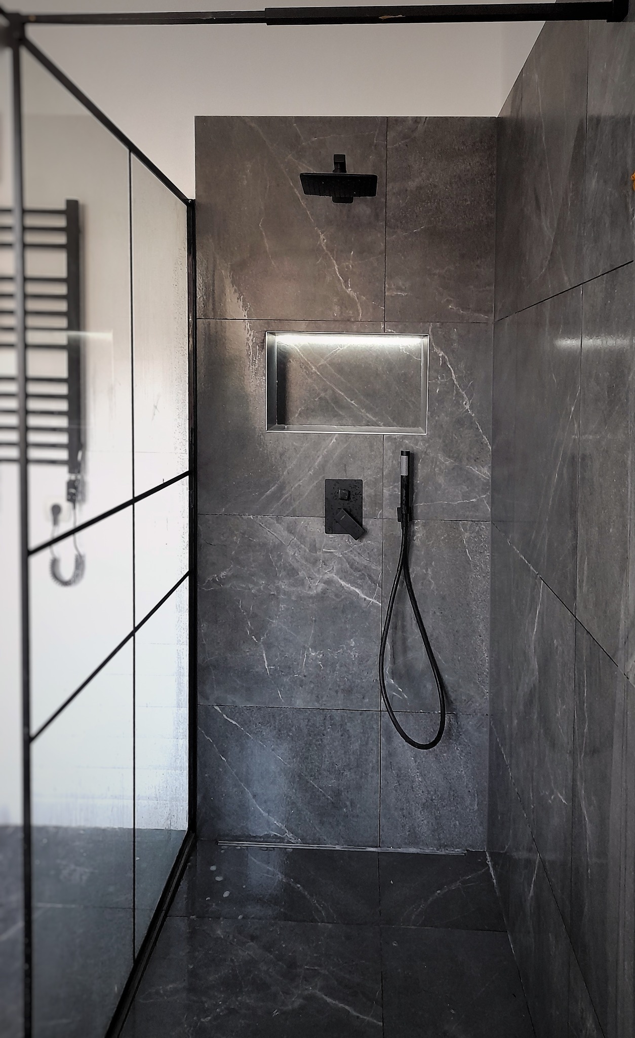 Nowoczesna kabina prysznicowa z szarymi płytkami imitującymi marmur, czarną armaturą i podświetlaną wnęką.