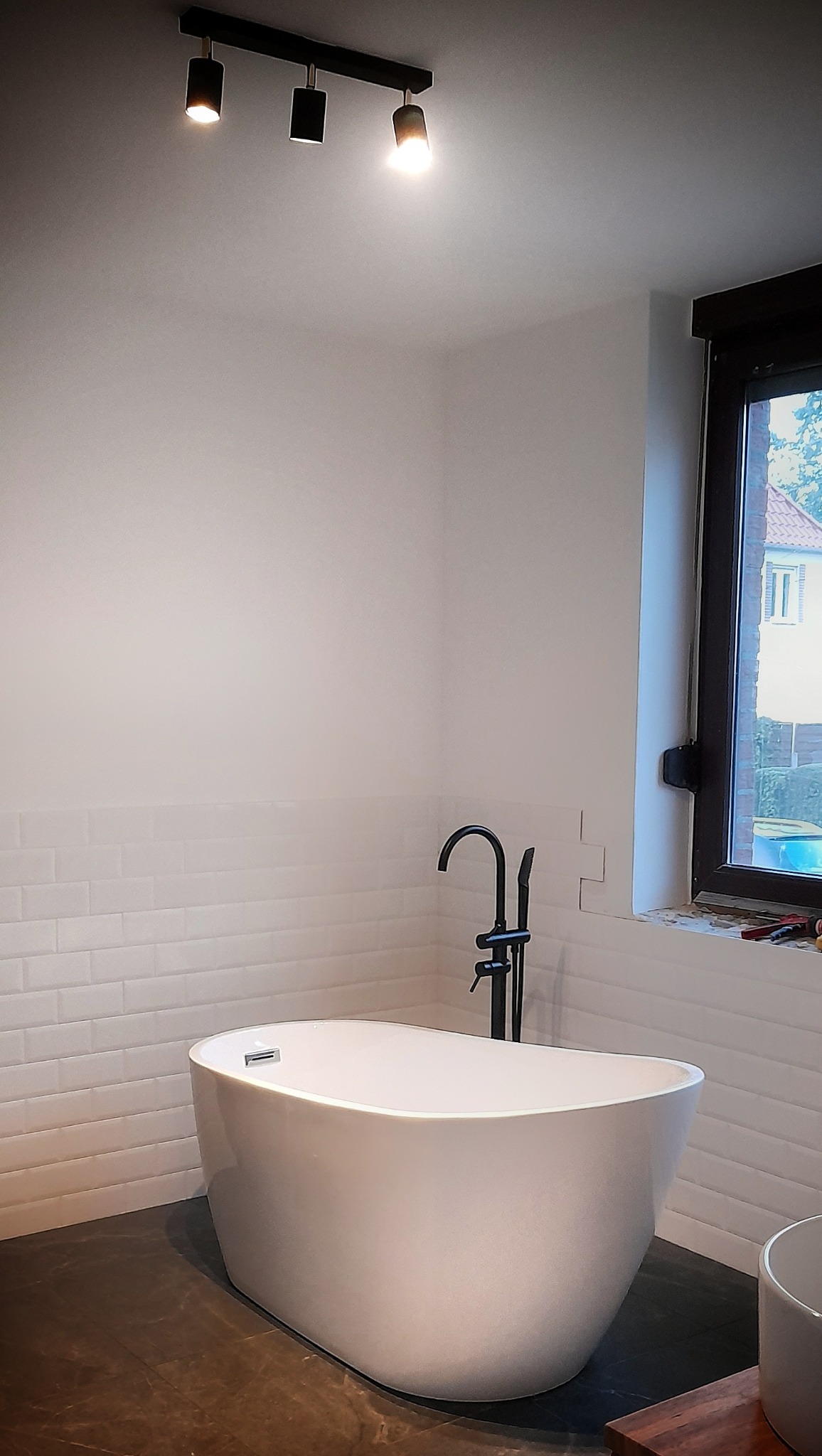 Biała, wolnostojąca wanna z czarną armaturą na tle ściany z białych płytek w kształcie cegiełek, oświetlona punktowym światłem