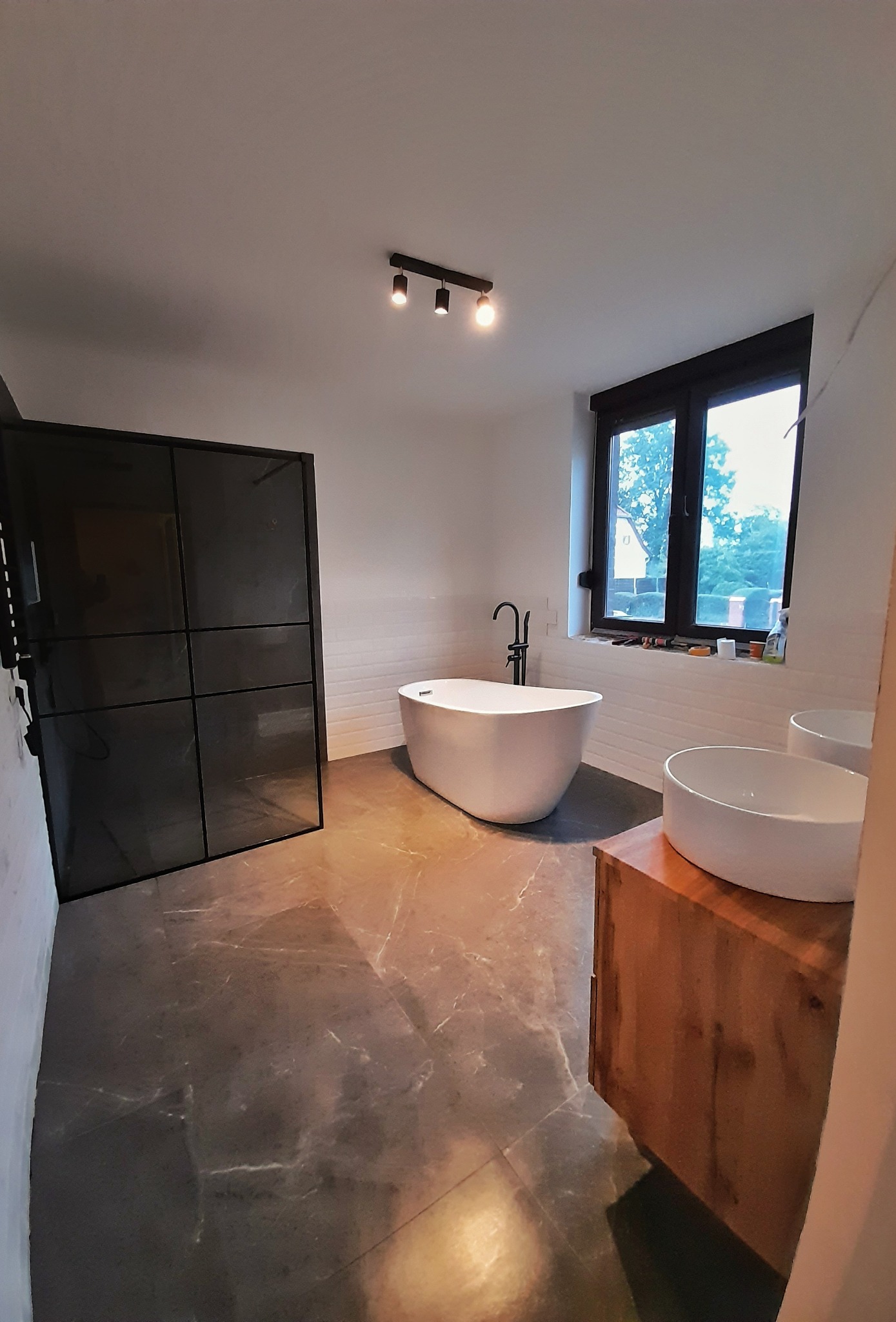 Nowoczesna łazienka z wolnostojącą wanną, kabiną prysznicową typu walk-in z czarnymi profilami, umywalką nablatową na drewnianej szafce i szarą marmurową podłogą.
