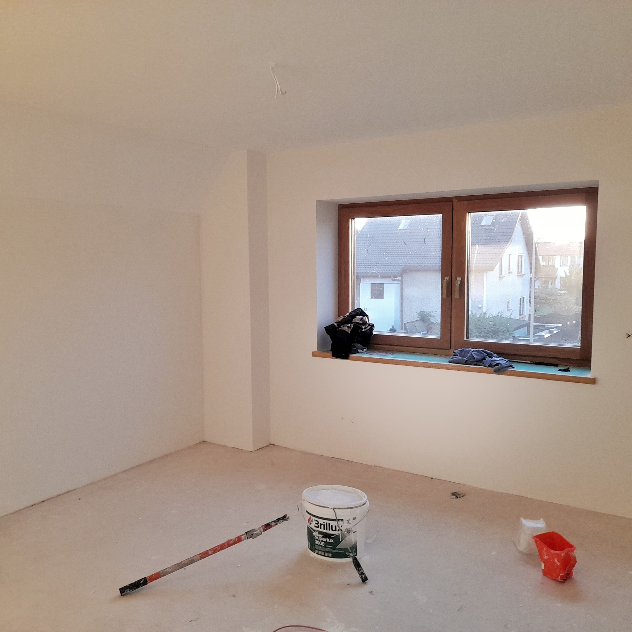 Przygotowanie do malowania: Puste, odświeżone pomieszczenie z oknem, wiadro farby Brillux Superlux 3000, wałek malarski z teleskopową rączką i kuweta na podłodze, gotowe do malowania ścian.
