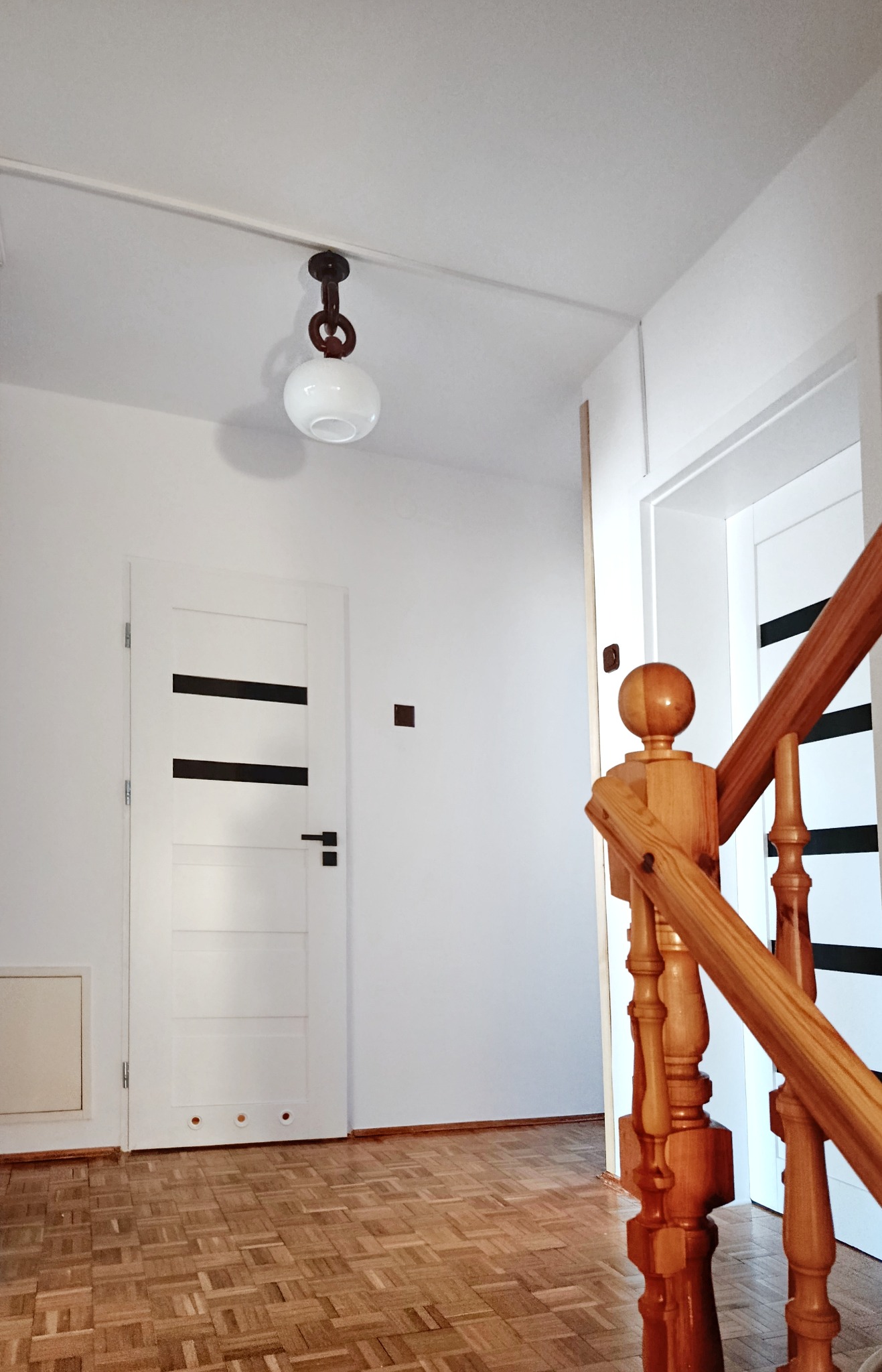 Białe wnętrze po malowaniu: ściany, drzwi z czarnymi pasami, drewniana podłoga parkietowa i balustrada schodów. Lampa sufitowa w stylu retro.