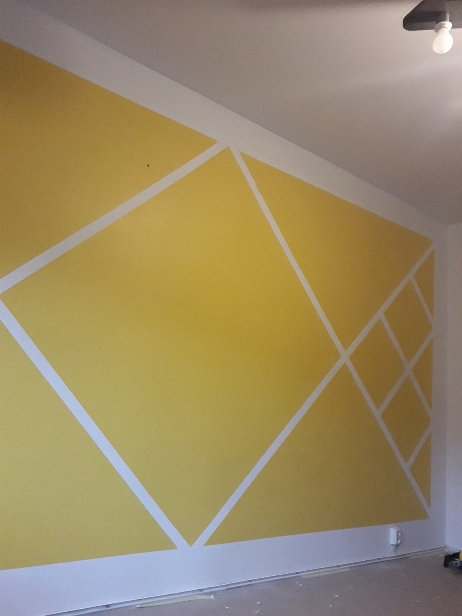 Żółta ściana z geometrycznym wzorem z białych linii, świeżo po malowaniu. Widoczne detale wykończenia i gniazdko elektryczne przy podłodze.
