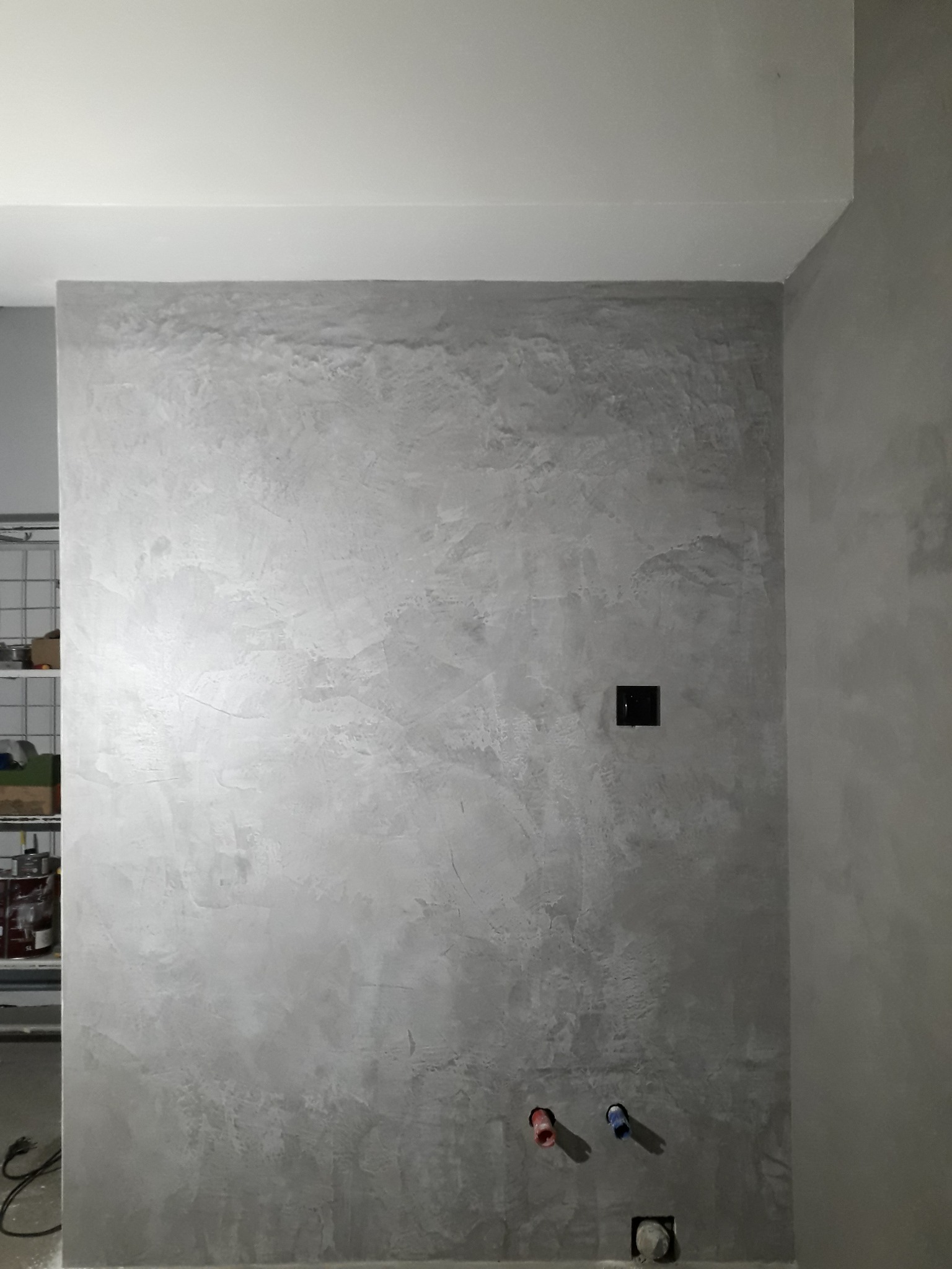 Ściana w łazience wykończona mikrocementem w odcieniach szarości, z widocznymi przyłączami wodnymi i gniazdkiem elektrycznym. Minimalistyczny design.