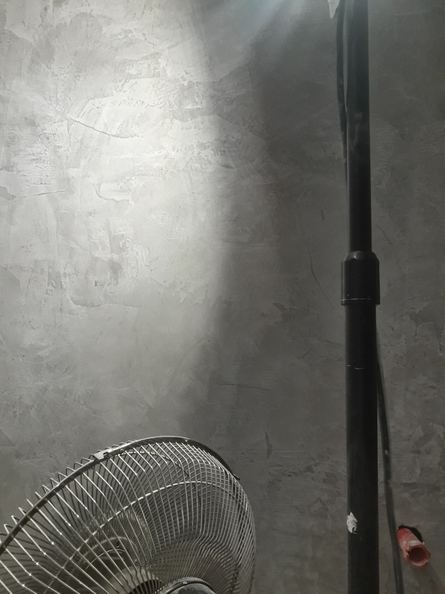 Szara ściana z efektem betonu, oświetlona z góry, z fragmentem wentylatora i czarną rurą. Minimalistyczne wykończenie wnętrza.