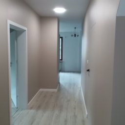 GRZEGORZ OKRAJEK SPEC-REM - Wykończony korytarz z panelami podłogowymi w odcieniach szarości, świeżo pomalowanymi ścianami w beżowym kolorze i nowoczesnym oświetleniem sufitowym.