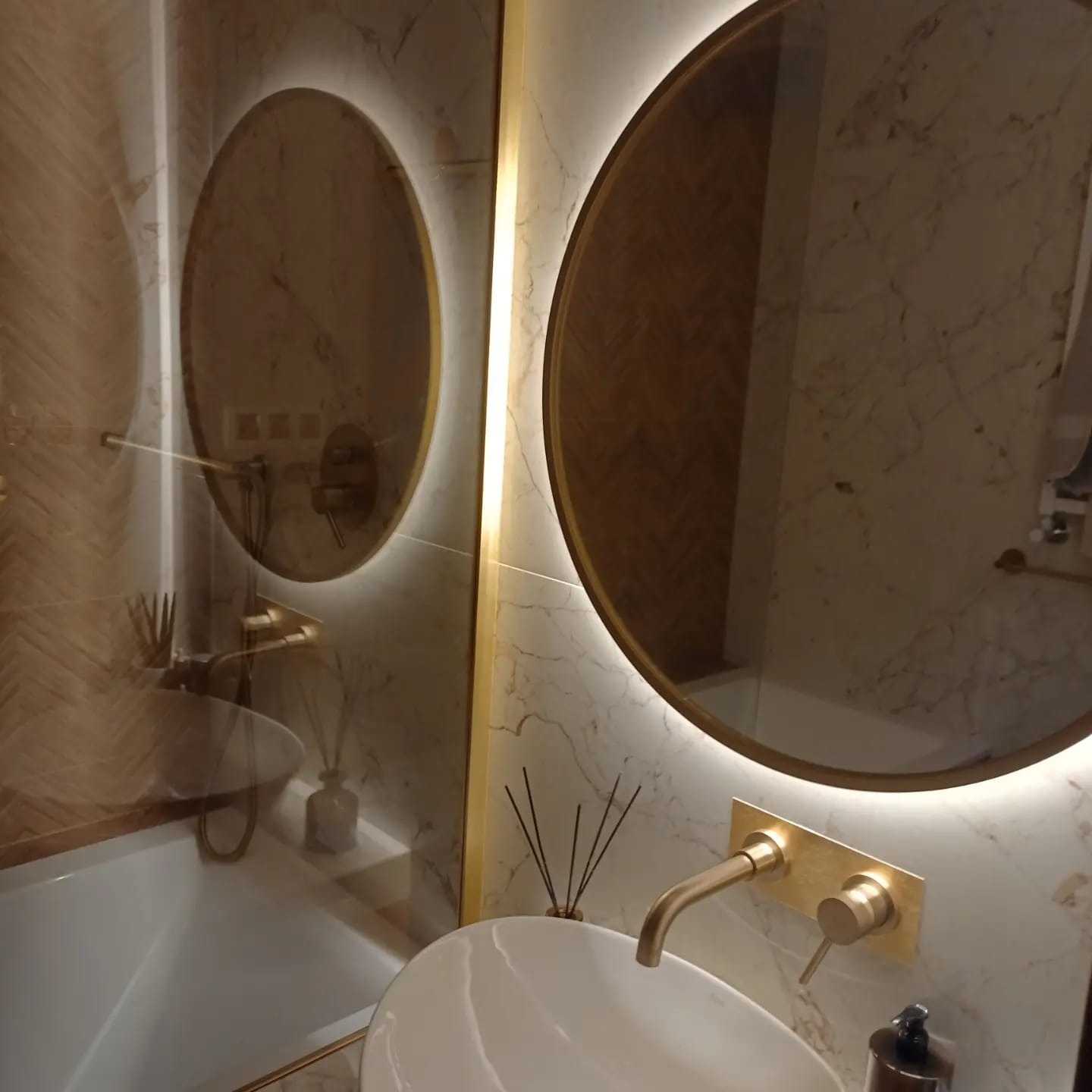 Nowoczesna łazienka z okrągłym lustrem podświetlanym LED, bateria umywalkowa w kolorze szczotkowanego złota i marmurowe płytki na ścianach.