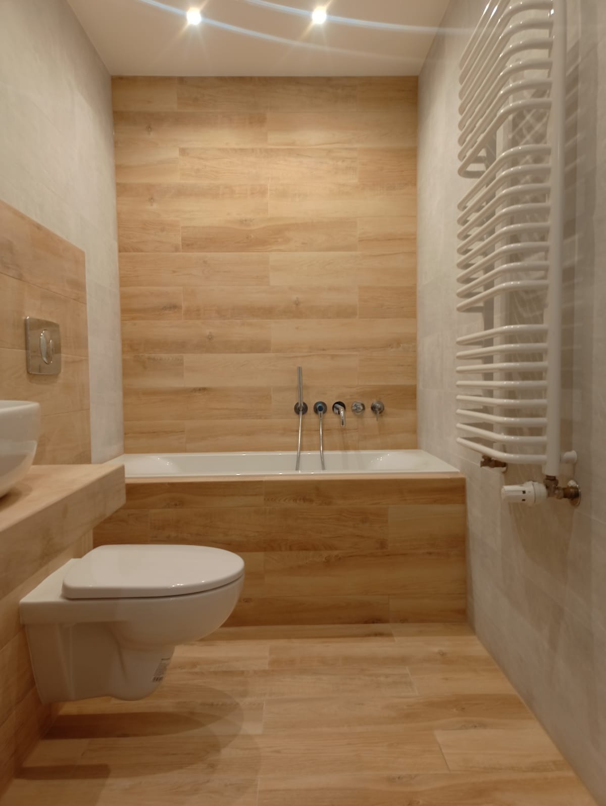 Nowoczesna łazienka wykończona w jasnym drewnie, z wiszącą toaletą, wanną obudowaną drewnem i białym grzejnikiem drabinkowym.