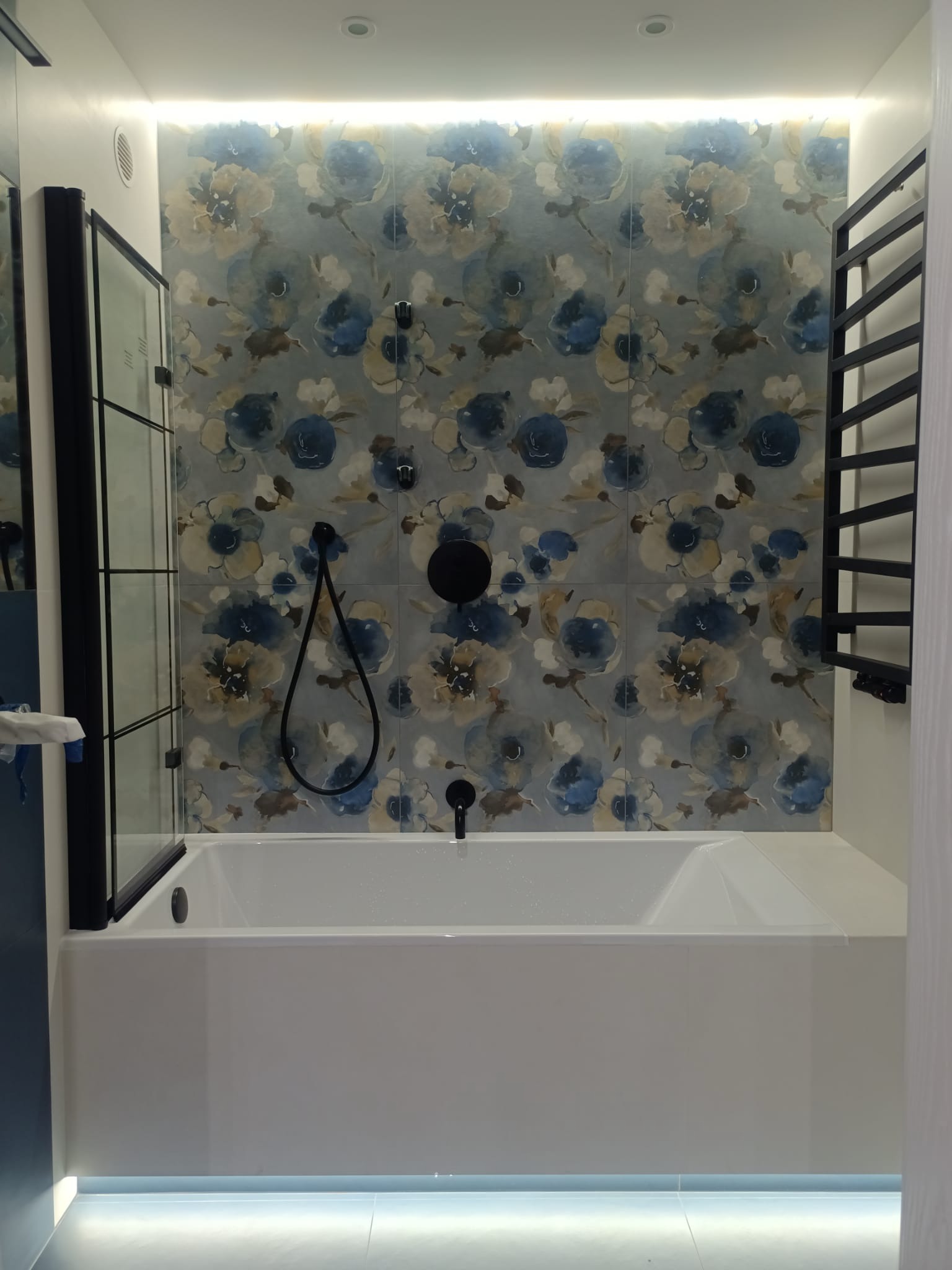 Nowoczesna łazienka z białą wanną, czarną armaturą i ścianą wyłożoną płytkami w kwiatowy wzór, oświetlenie LED pod wanną i nad płytkami.