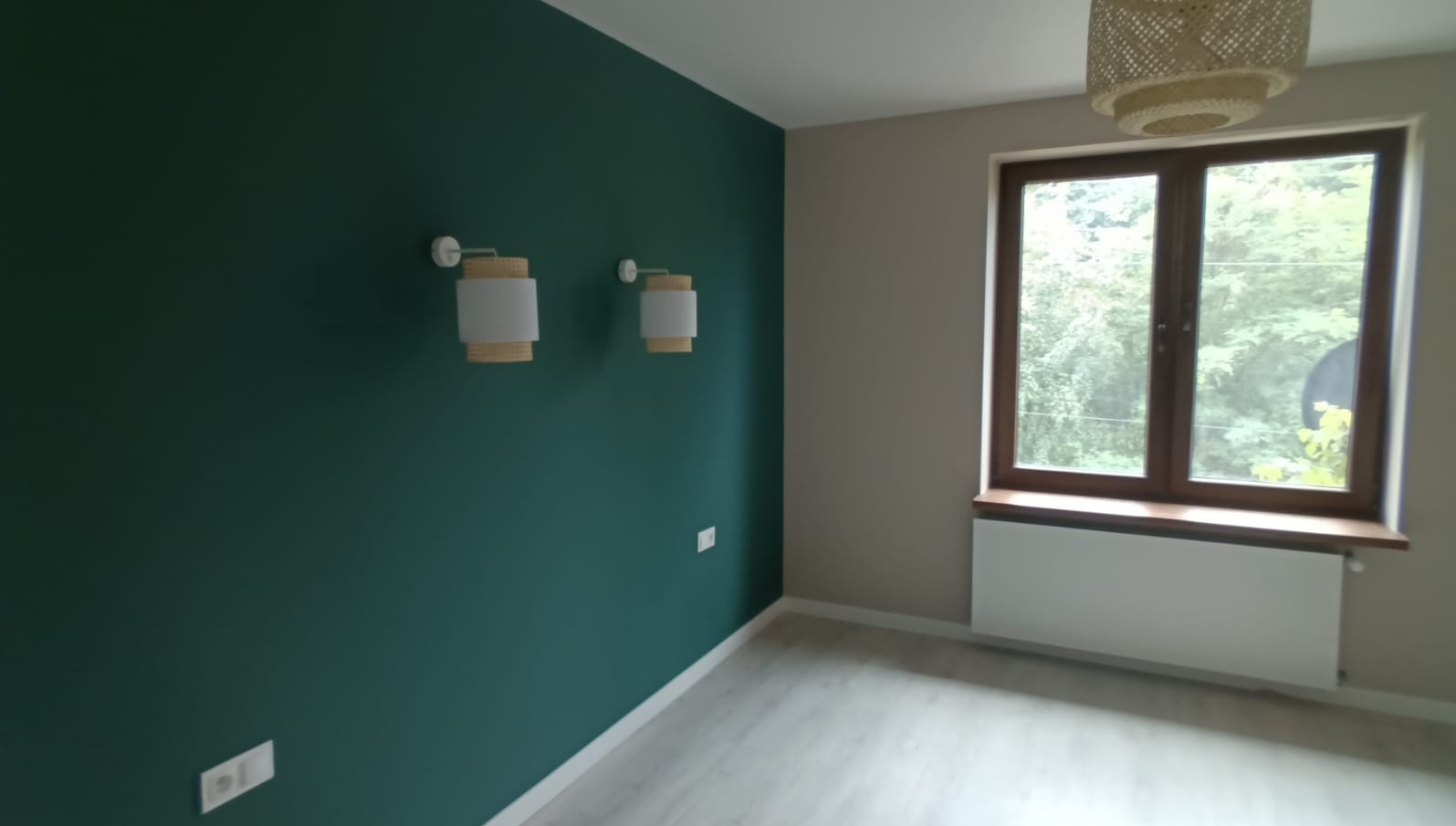 Pokój po remoncie: ściana pomalowana na ciemnozielono z dwoma kinkietami, jasna podłoga i okno z widokiem na zieleń.