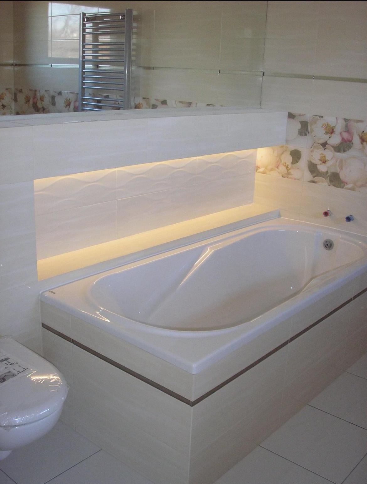 Wykończona łazienka z wanną obudowaną płytkami, podświetlanym panelem dekoracyjnym i toaletą w folii ochronnej.