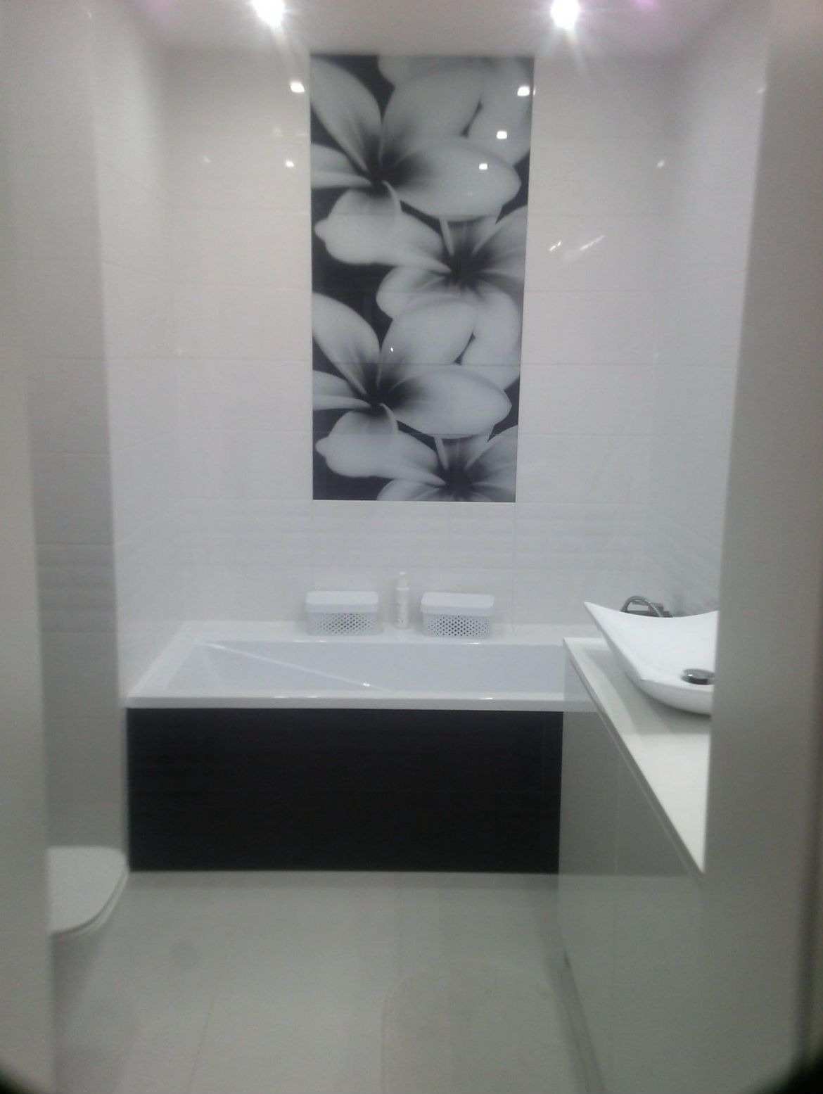 Nowoczesna łazienka z białą armaturą, czarnym panelem pod wanną i dekoracyjną płytką z motywem kwiatowym na ścianie.