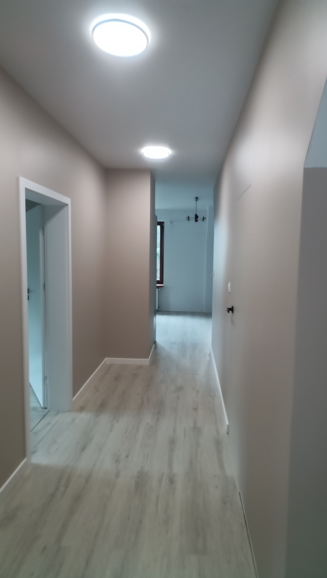 Wykończony korytarz z panelami podłogowymi w odcieniach szarości, świeżo pomalowanymi ścianami w beżowym kolorze i nowoczesnym oświetleniem sufitowym.