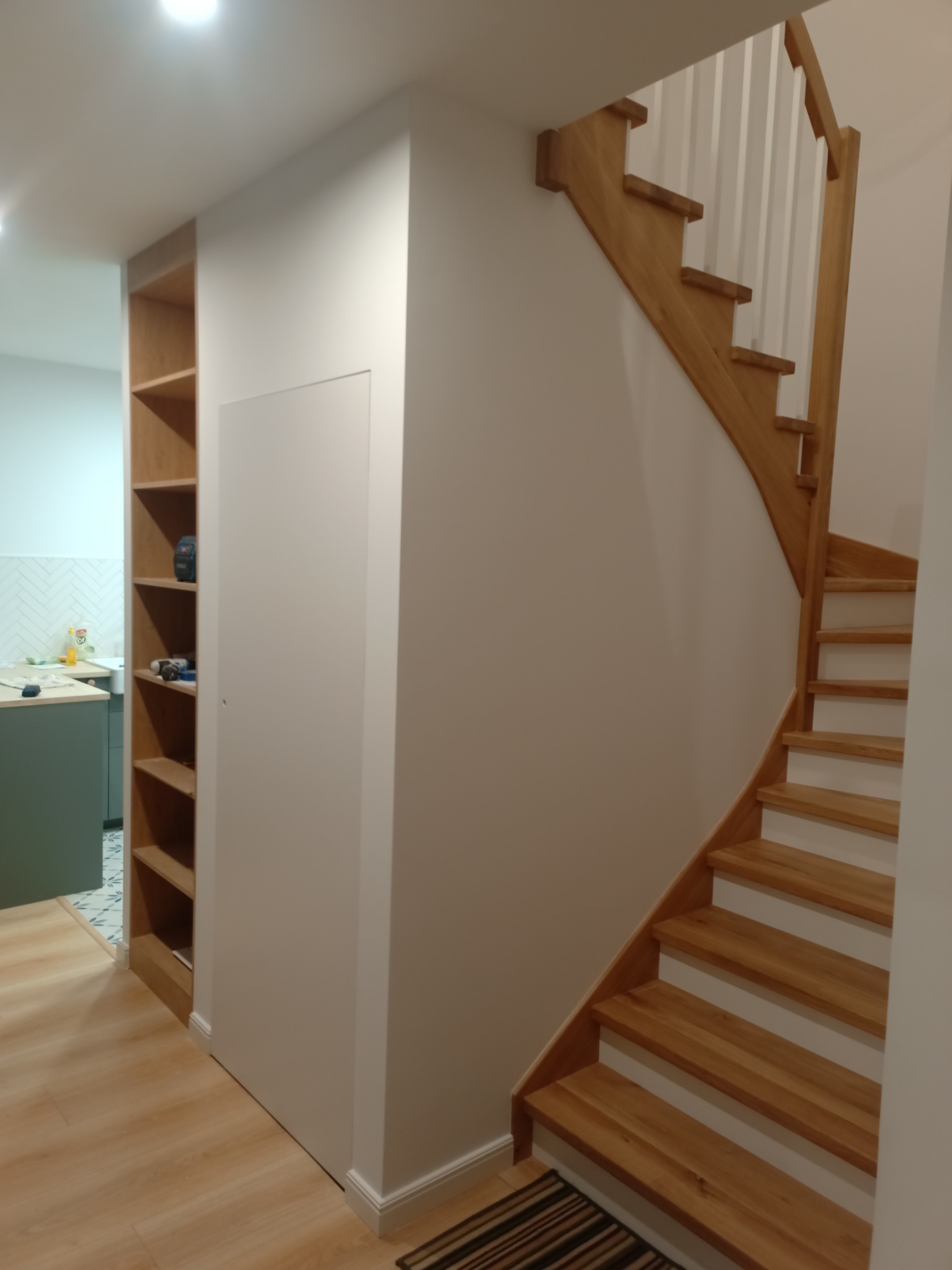 Jasne drewniane schody z białymi podstopnicami, zabudowa regałowa z jasnego drewna i białe ściany w nowoczesnym wnętrzu.