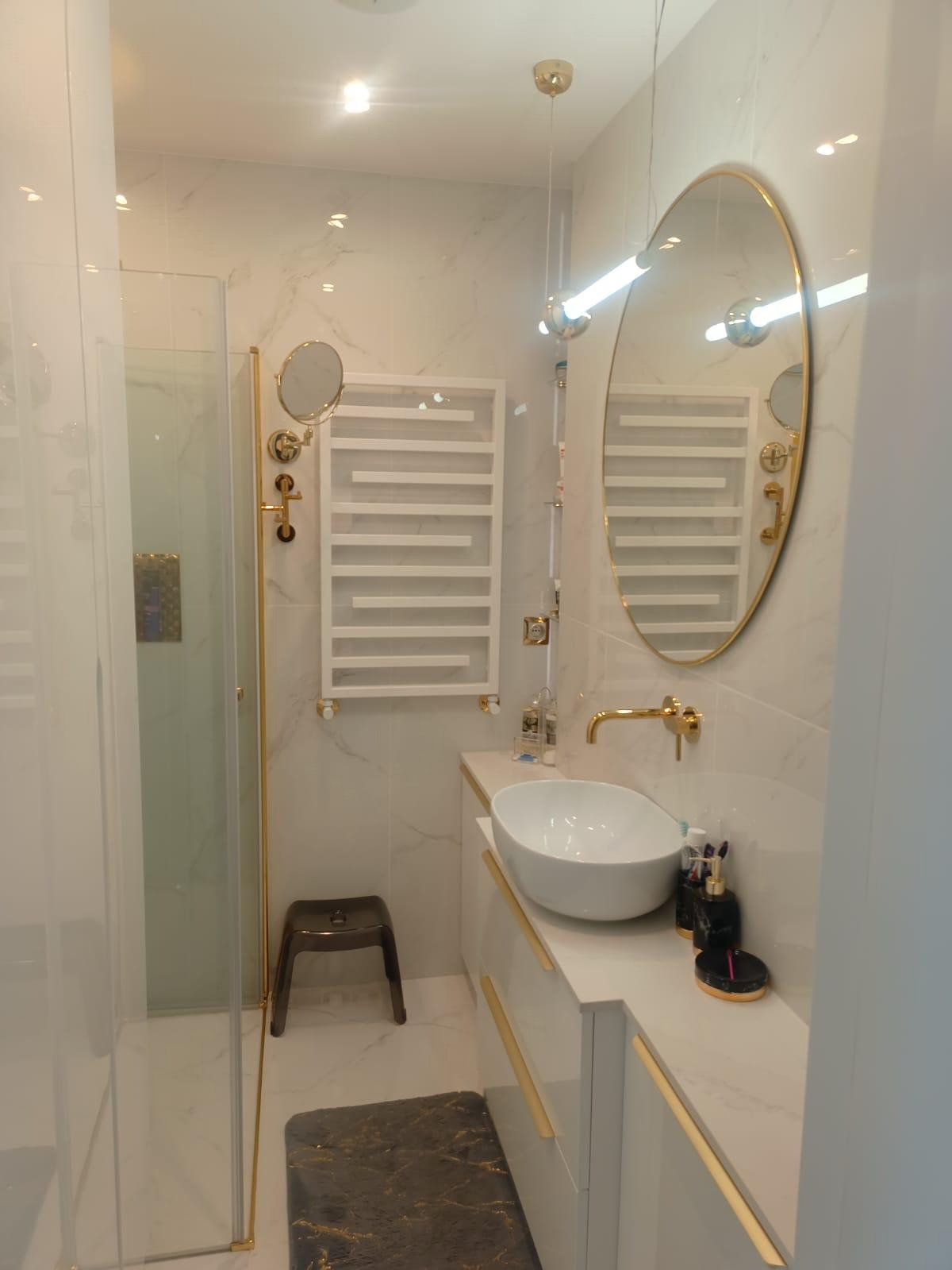 Nowoczesna łazienka z marmurowymi płytkami, owalnym lustrem w złotej ramie, białą umywalką nablatową i grzejnikiem drabinkowym w minimalistycznym stylu.