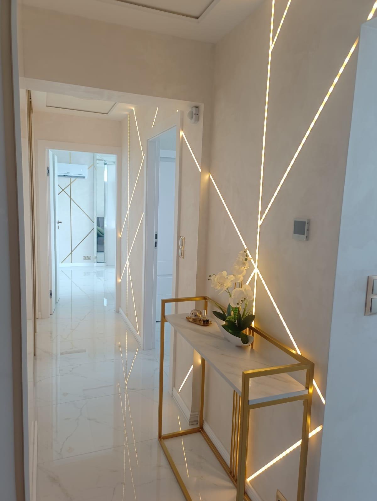 Nowoczesny korytarz z marmurową podłogą, ścianami zdobionymi geometrycznymi liniami świetlnymi i konsolą z blatem imitującym marmur oraz złotą ramą, na której stoi wazon z białymi kwiatami.