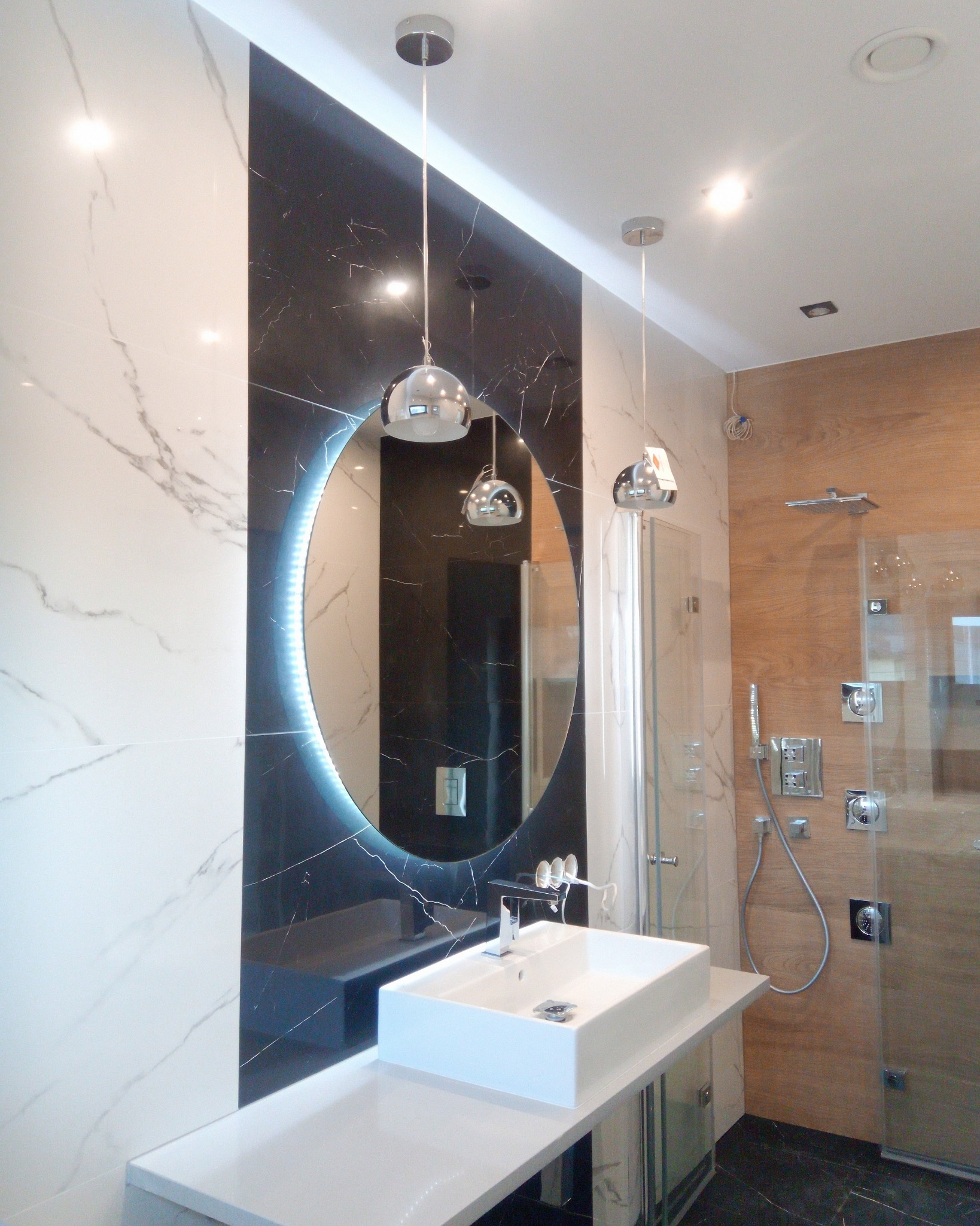 Nowoczesna łazienka z owalnym lustrem podświetlanym LED, białą umywalką nablatową i kabiną prysznicową z drewnopodobnymi panelami.