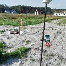 GEO-GRAF USŁUGI GEODEZYJNE - Pomiarowy odbiornik GNSS na tyczce, ustawiony na działce budowlanej z widocznym wykopem studzienki kanalizacyjnej i powstającymi domami w tle.