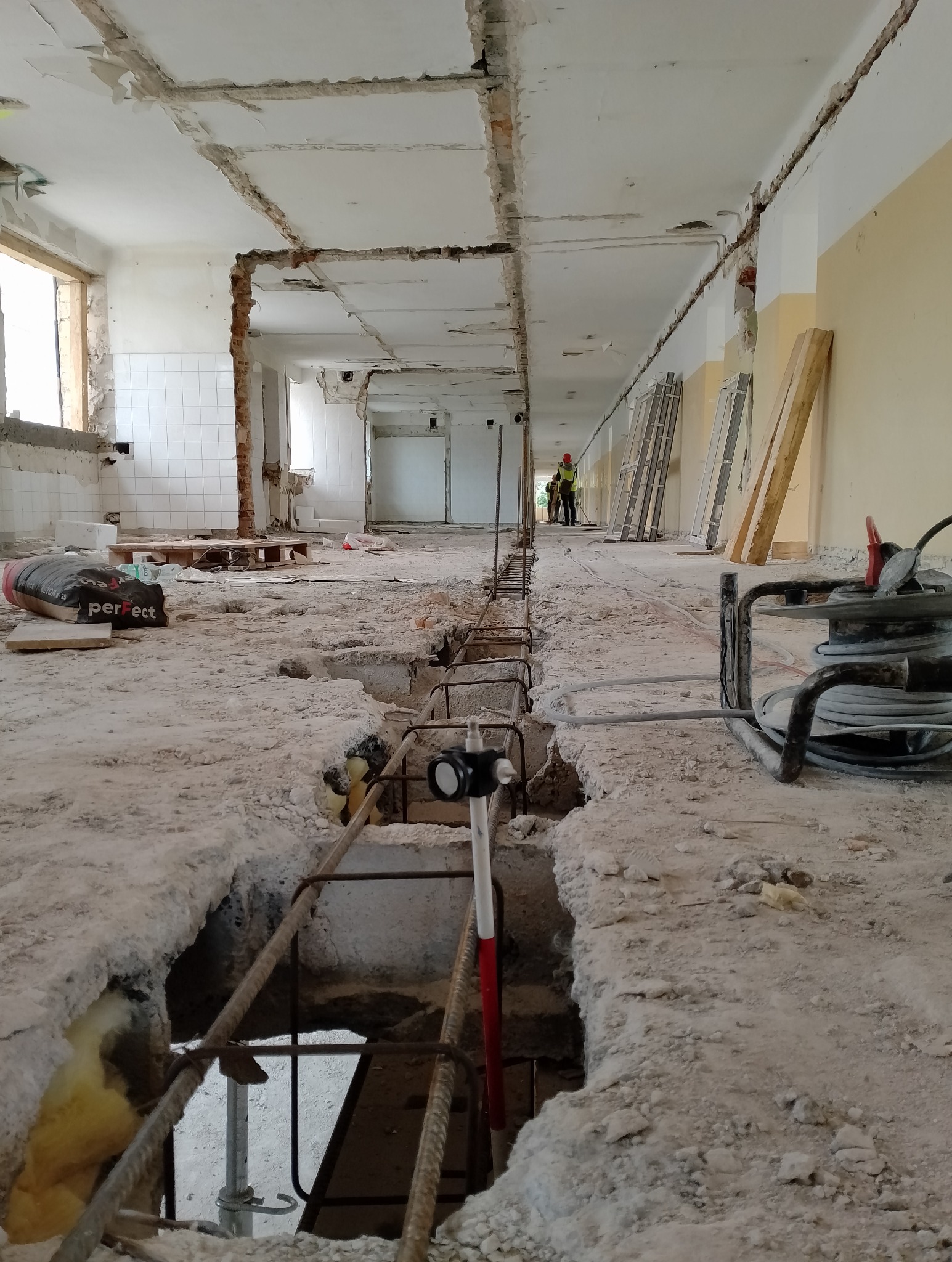 Wnętrze budynku w trakcie remontu: wykute bruzdy w podłodze z widocznym zbrojeniem, ekipa budowlana w oddali, worki z zaprawą budowlaną i kable elektryczne w tle.