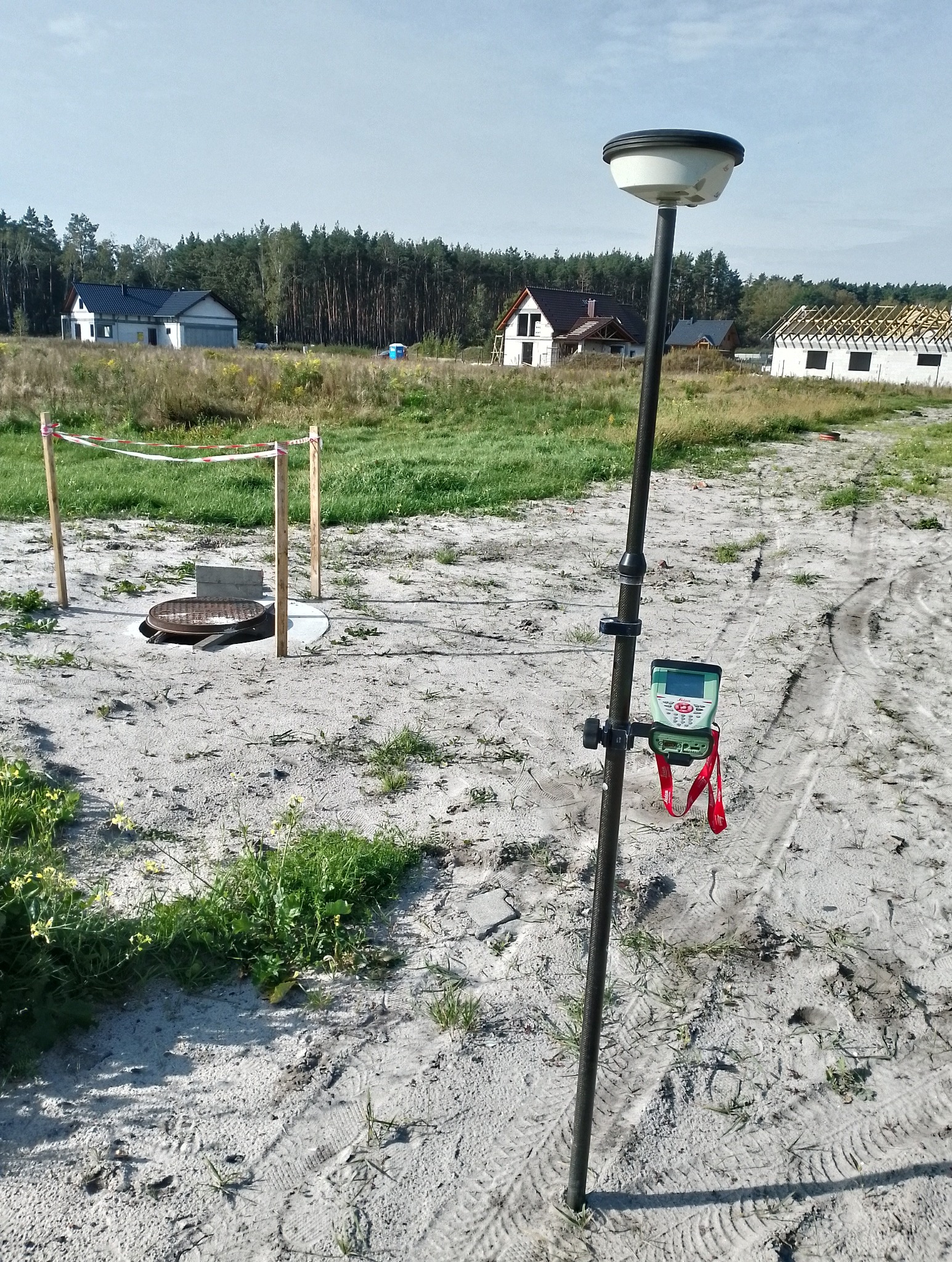 Pomiarowy odbiornik GNSS na tyczce, ustawiony na działce budowlanej z widocznym wykopem studzienki kanalizacyjnej i powstającymi domami w tle.