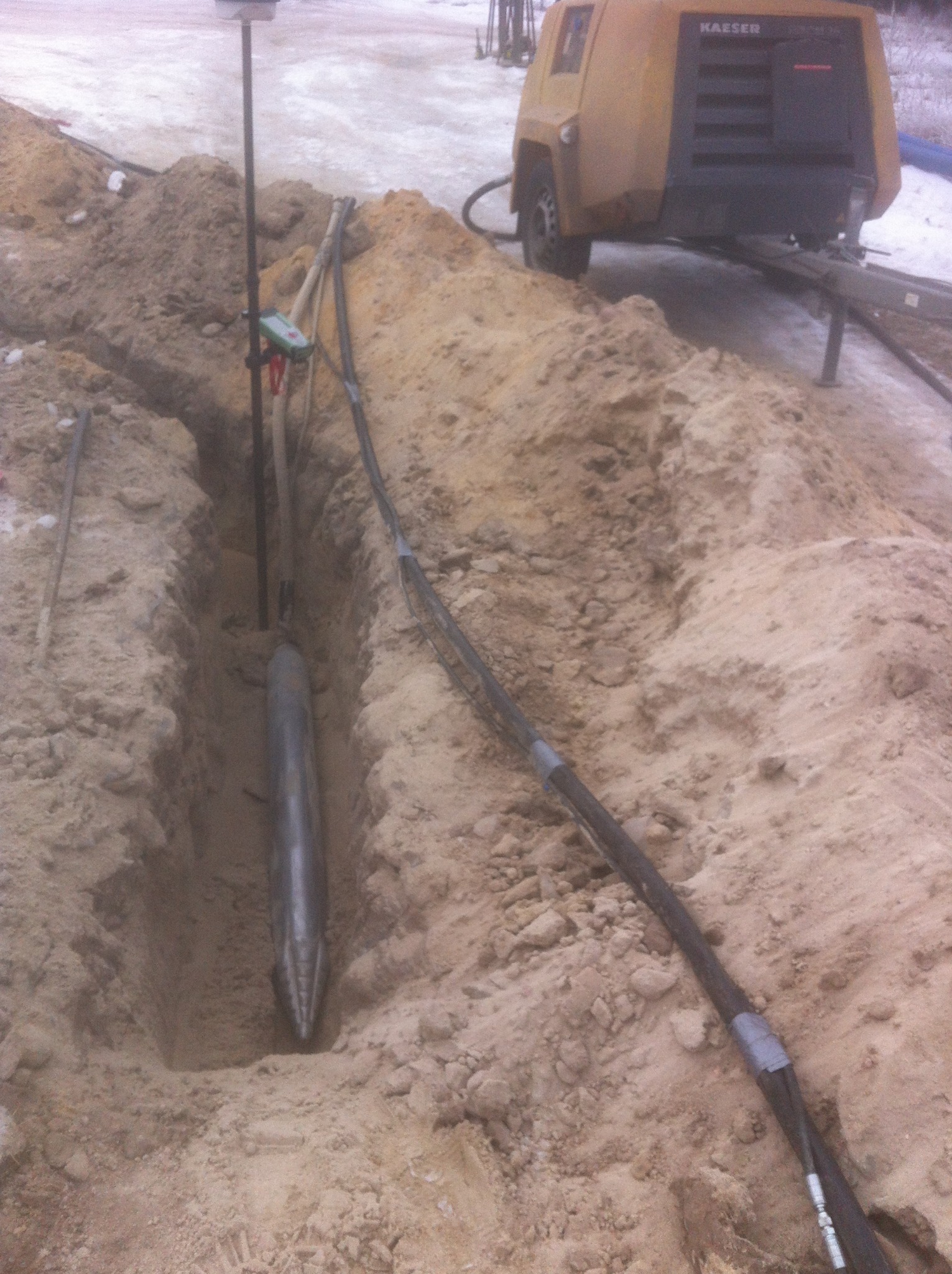 Wykop z umieszczoną w nim sondą wiertniczą, obok widoczny agregat sprężarkowy Kaeser i tyczka geodezyjna z odbiornikiem GPS.