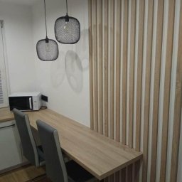 NA WYMIAR Agnieszka Janeczko - Nowoczesna aranżacja wnętrza z drewnianym blatem, dwoma krzesłami i dekoracyjną ścianą z drewnianych listew. Nad blatem wiszą dwie czarne lampy.