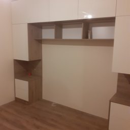 NA WYMIAR Agnieszka Janeczko - Biało-drewniana szafa wnękowa z półkami i szufladami, oświetlona od góry, na tle drewnianej podłogi. Minimalistyczny design, zabudowa od podłogi do sufitu.