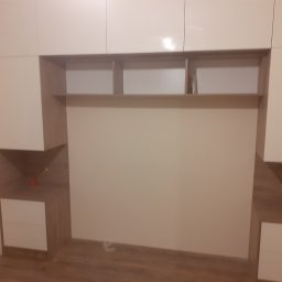 NA WYMIAR Agnieszka Janeczko - Jasna zabudowa meblowa z szafkami górnymi i szafkami nocnymi po bokach, z otwartymi półkami nad przestrzenią łóżka. Fronty szafek lakierowane na wysoki połysk.