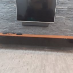 NA WYMIAR Agnieszka Janeczko - Nowoczesna szafka RTV z czarnymi frontami i drewnianym blatem, zawieszona pod telewizorem na tle ściany z szarej cegły dekoracyjnej. Minimalistyczny design.