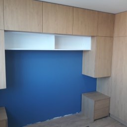 NA WYMIAR Agnieszka Janeczko - Jasna szafa wnękowa z drewnianymi frontami i białymi półkami, na tle niebieskiej ściany. Widoczna szafka nocna i lustrzane drzwi. Minimalistyczny design.