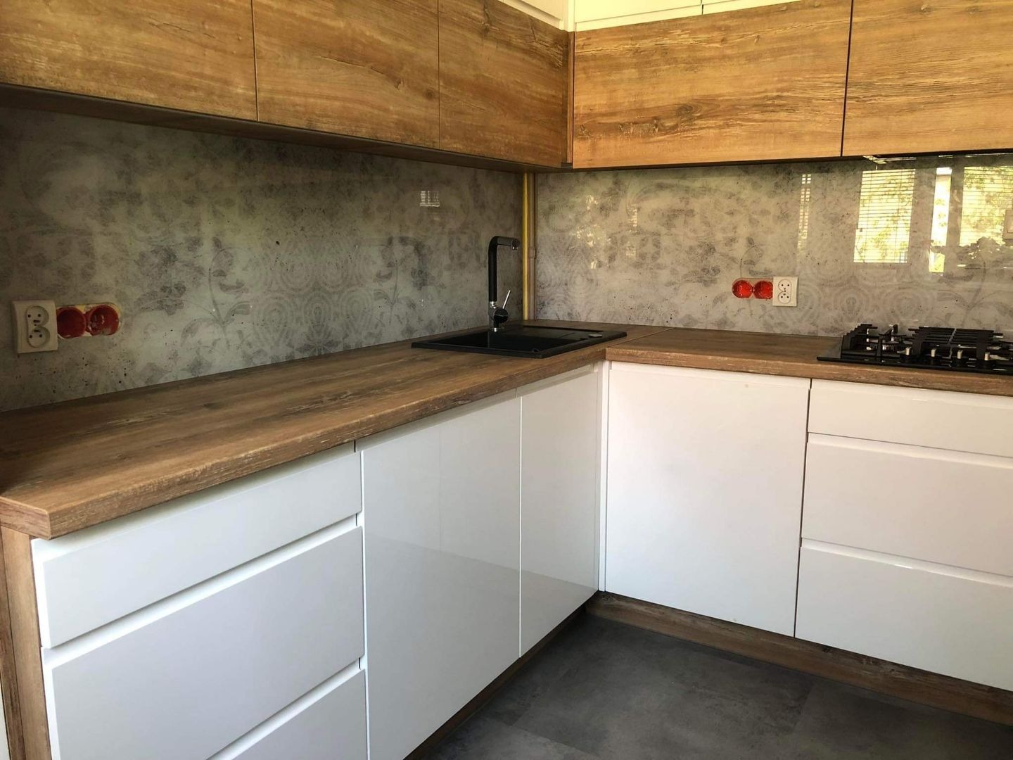 Nowoczesna kuchnia narożna z białymi, lakierowanymi frontami i drewnianym blatem. Na ścianie dekoracyjny panel z subtelnym wzorem. Widoczne gniazdka i płyta grzewcza.