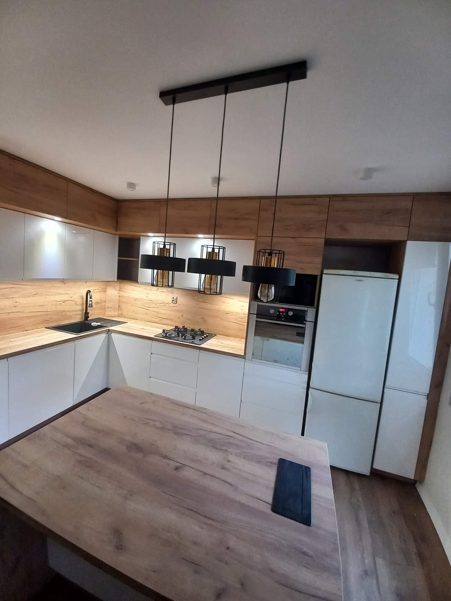 Nowoczesna kuchnia na wymiar z białymi frontami i drewnianymi akcentami, oświetlona wiszącymi lampami. Na pierwszym planie drewniany stół z czarnym gniazdkiem.