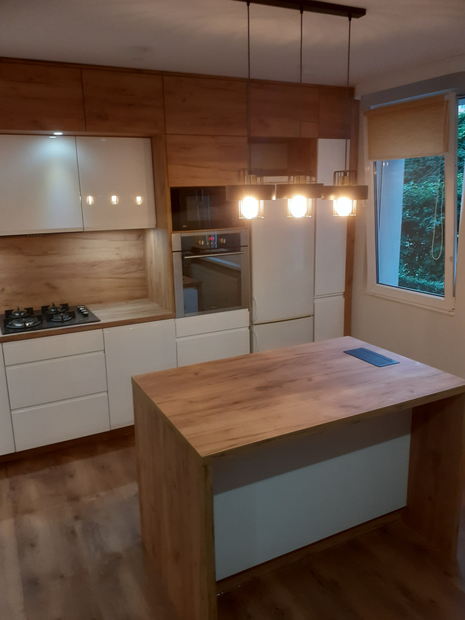 Nowoczesna kuchnia z wyspą: białe fronty, drewniane akcenty, oświetlenie wiszące. Widok na okno z roletą i zielenią. Uwagę przyciąga drewniany blat wyspy.