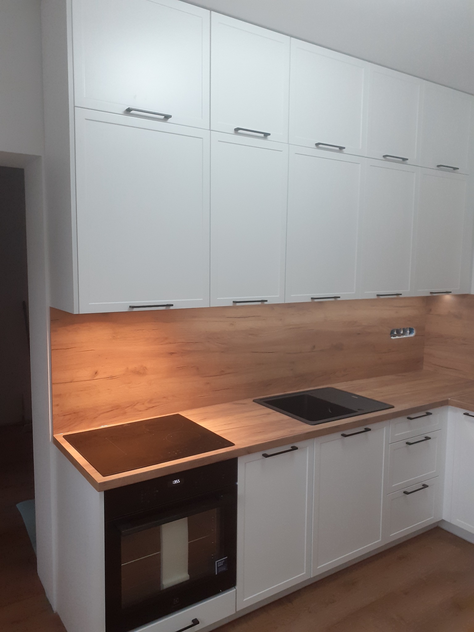 Biała kuchnia z drewnianym blatem i czarnymi uchwytami, płyta indukcyjna i piekarnik w zabudowie, minimalistyczny design, oświetlenie podszafkowe.