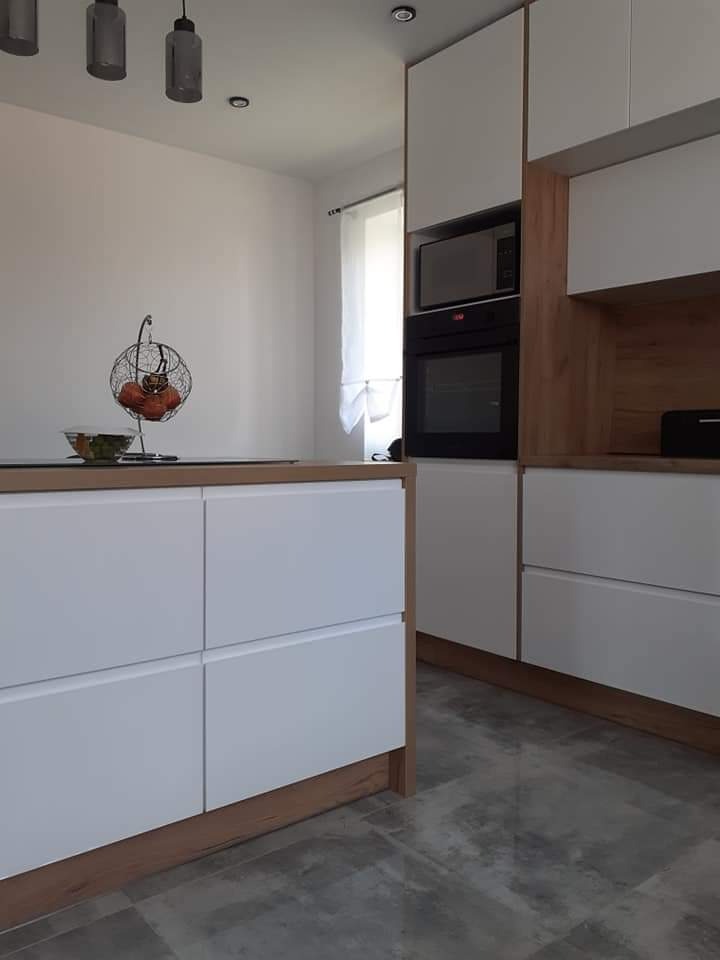 Nowoczesna kuchnia z białymi frontami i drewnianymi akcentami, widok na wyspę kuchenną z owocami w metalowym koszu, szare płytki podłogowe i oświetlenie sufitowe.