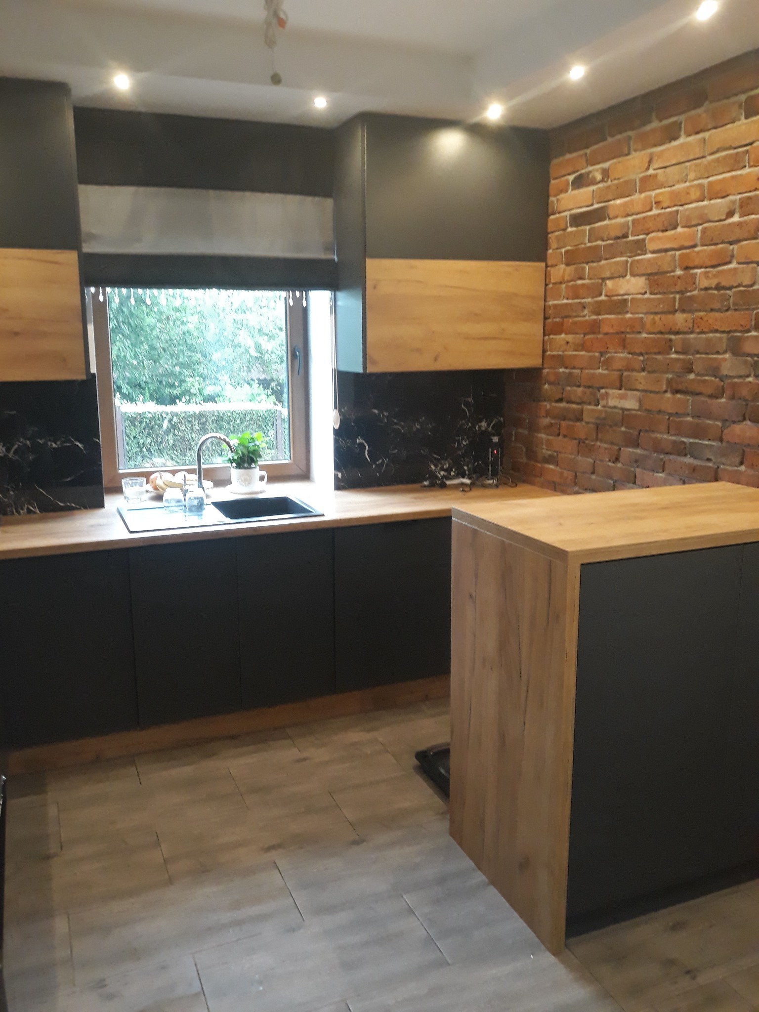 Nowoczesna kuchnia z czarnymi szafkami i drewnianymi blatami, ceglana ściana w tle. Widok na okno z roślinami. Oświetlenie punktowe.